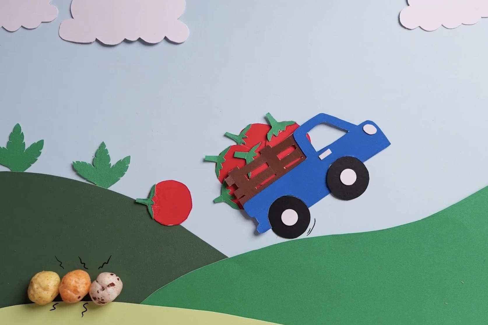 Hand-cut paper set for a stop motion frame: a blue truck carrying tomatoes over green hills