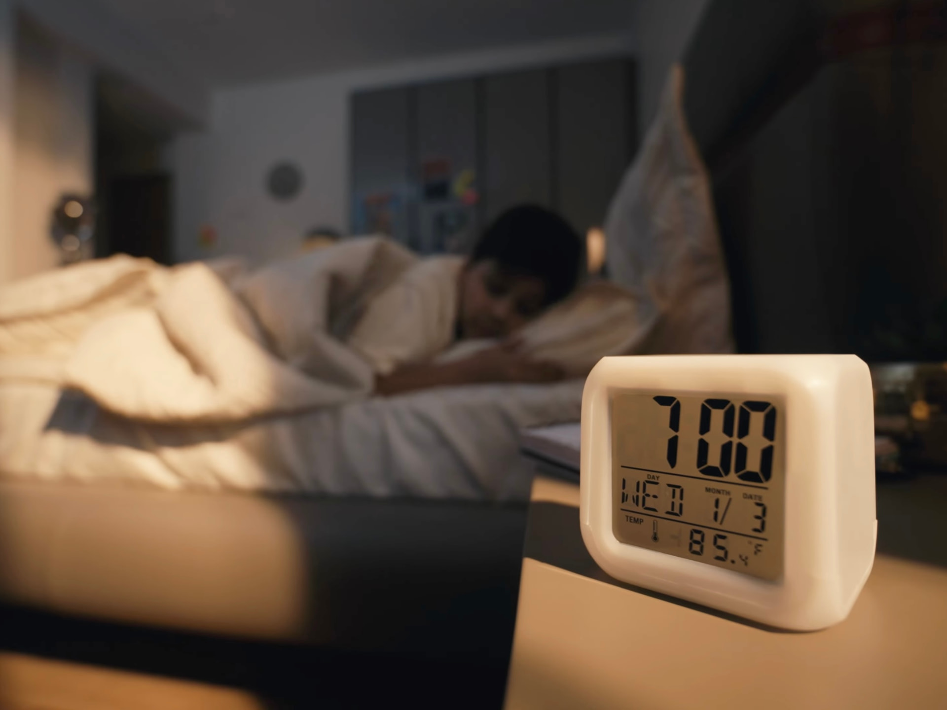 Macro film frame of a digital alarm clock reading 7:00 with a child asleep behind it