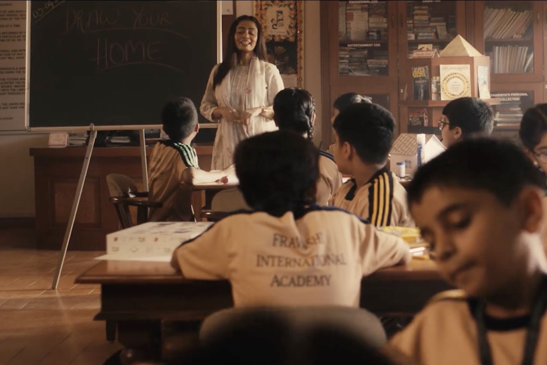 Ad film frame of a teacher in a classroom with children drawing their homes